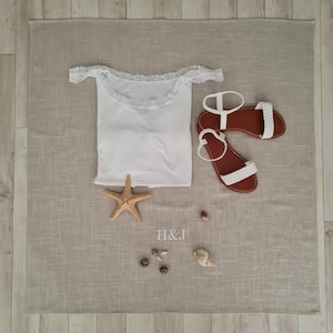 Può includere: Un top bianco con spalle scoperte e bordo in pizzo, un paio di sandali bianchi con suola marrone, una stella marina e diverse piccole conchiglie disposte su uno sfondo di tessuto grigio chiaro testurizzato con le lettere H&J ricamate al centro.