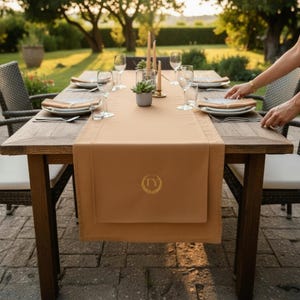 Peut inclure: Une table à manger en bois dressée pour un repas en plein air. La table est recouverte d'un chemin de table beige et garnie d'assiettes, de couverts et de verres à vin. Une petite plante succulente et des bougies sont au centre. Le chemin de table porte un emblème doré avec les lettres "TY".