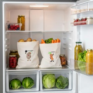 May include: Two off-white cotton produce bags sitting on a refrigerator shelf. The left bag features a cartoon potato illustration with the text POTATOES. The right bag features a cartoon carrot illustration with the text CARROT.