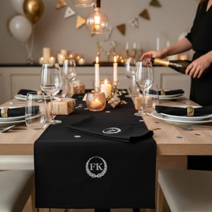 Peut inclure: Une table de salle à manger décorée pour une célébration, avec un chemin de table noir et des serviettes assorties brodées de détails blancs. La table est ornée de verres, de bougies et d'éléments dorés. Une personne verse du champagne en arrière-plan.