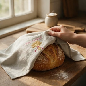 May include: A light beige linen kitchen towel featuring an embroidered golden wheat sheaf tied with a pink ribbon and the text Kimie Sourdough in pink thread, draped over a round loaf of crusty sourdough bread on a wooden surface.