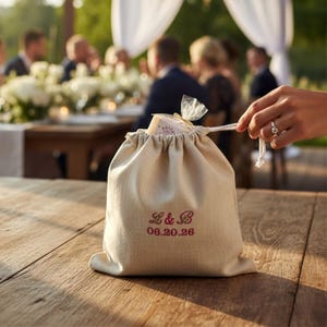 Op de afbeelding: Een beige stoffen buidel met trekkoord voorzien van roze geborduurde letters L & B en de datum 06.20.26. De tas is aan de bovenkant dichtgetrokken met een wit koord en bevat een klein item verpakt in doorzichtig plastic, zichtbaar bij de opening.