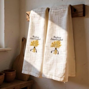 Puede incluir: Dos paños de cocina blanquecinos colgados de un estante de madera. Cada paño presenta un diseño de trigo bordado en oro y negro, con el texto "Andreas Sauerteigbrot" encima. Los paños tienen un aspecto texturizado.