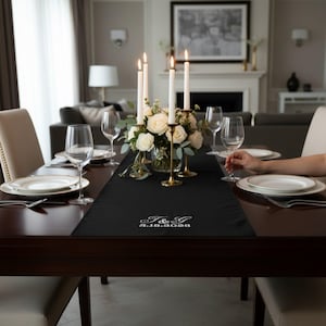 Peut inclure: Un chemin de table noir avec une broderie blanche indiquant T & G 8.18.2026. Il est disposé sur une table à manger en bois foncé avec des assiettes blanches, des verres à vin transparents et un centre de table de roses blanches et quatre bougies.