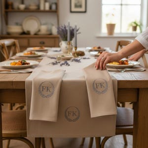 May include: A dining table set for a meal, with a beige tablecloth and matching napkins. Each napkin and the tablecloth feature a blue circular design with the initials "FK" and a laurel wreath. Plates of food and decorative items are arranged on the table.