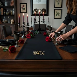 Peut inclure: Une table de salle à manger formelle avec un chemin de table noir brodé de lettres dorées, des roses rouges, des gobelets noirs et des bougies bordeaux allumées dans des candélabres noirs ornés. Une personne arrange la table.