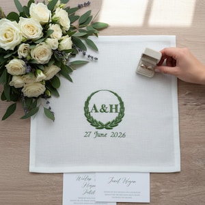 Può includere: Un tovagliolo di lino bianco con ricamo verde di una corona d'alloro che circonda le lettere A&H e la data 27 giugno 2026. A sinistra si trova un bouquet di rose bianche e lavanda, con un piccolo portafedi posizionato sul tessuto.