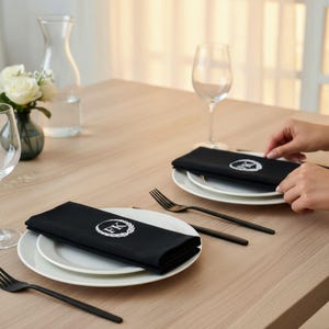 May include: Black cloth napkins featuring a white embroidered circular laurel wreath design with the letters FK in the center. The napkins are folded and placed on white ceramic plates, accompanied by black metal forks on a light wood table.