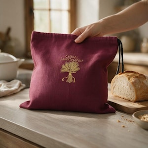 May include: A deep burgundy fabric drawstring bag featuring an embroidered gold wheat sheaf graphic and the text "Celines Sauerteigbrot" in gold thread. The bag has a dark drawstring cord and is designed for storing bread.