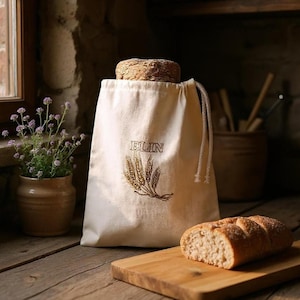 May include: A cream-coloured canvas bread bag with the word "ELIN" and a wheat stalk design embroidered in brown. A loaf of bread sits on a wooden chopping board. A small potted plant and a loaf of bread are also visible.