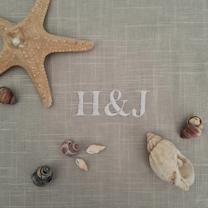 Op de afbeelding: Een lichtbeige linnen stof met de geborduurde witte letters H & J in het midden. De stof is versierd met een grote zandkleurige zeester in de linkerbovenhoek en verschillende kleine, gevarieerde schelpen verspreid over het oppervlak.