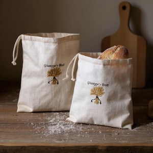 Può includere: Due sacchi per il pane in tela bianco sporco con chiusure a coulisse. Un sacco contiene una pagnotta di pane. Ogni sacco ha il testo ricamato "Giuseppe's Brot" e un disegno di spiga di grano. I sacchi sono su una superficie di legno.