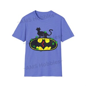 May include: A light blue t-shirt with a black cat sitting on a yellow Batman logo with green vines. The logo has a red diamond in the center.