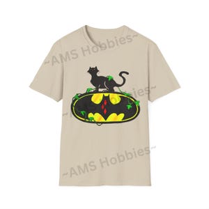 May include: A beige t-shirt with a black cat sitting on a yellow and black Batman logo with green vines. The logo has a red diamond in the center.