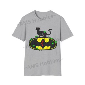 May include: A gray t-shirt with a black cat sitting on a yellow and black Batman logo with green vines. The logo has a red diamond in the center.