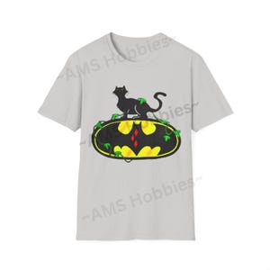 May include: A gray t-shirt with a black cat sitting on top of a yellow and black Batman logo with green vines. The logo has a red diamond in the center.