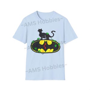 May include: Light blue t-shirt with a black cat sitting on top of a yellow Batman logo with green vines. The text "AMS Hobbies" is printed on the shirt.
