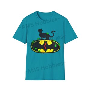 May include: A turquoise blue t-shirt with a black cat sitting on top of a yellow Batman logo with green vines. The text "AMS Hobbies" is printed on the shirt.
