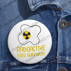 May include: A white button with a yellow fried egg design and a yellow radiation symbol inside. The text "RADIOACTIVE EGG SURVIVOR" is written in black below the egg.