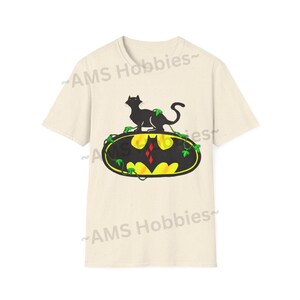 May include: A beige t-shirt with a black cat sitting on top of a yellow Batman logo with green vines. The logo has a red diamond in the center. The text "AMS Hobbies" is printed on the shirt.