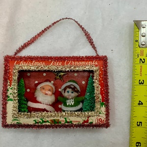 May include: A vintage Christmas tree ornament featuring a small Santa Claus and elf figurine in a red and white frame with a gold glitter border. The ornament is decorated with red and white polka dots and a small green Christmas tree. The text "Christmas Tree Ornaments" and "19&cent;" are visible on the ornament.