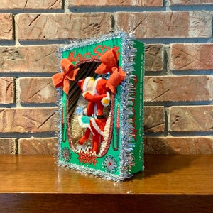 Op de afbeelding: Een vintage kerstboomversiering met een rood-witte kerstmanfiguur in een groen-witte doos met zilveren tinselrand. De doos heeft de tekst "Motor Star" en "A Trim of the Tree's Product by Delta Ornaments, Inc."