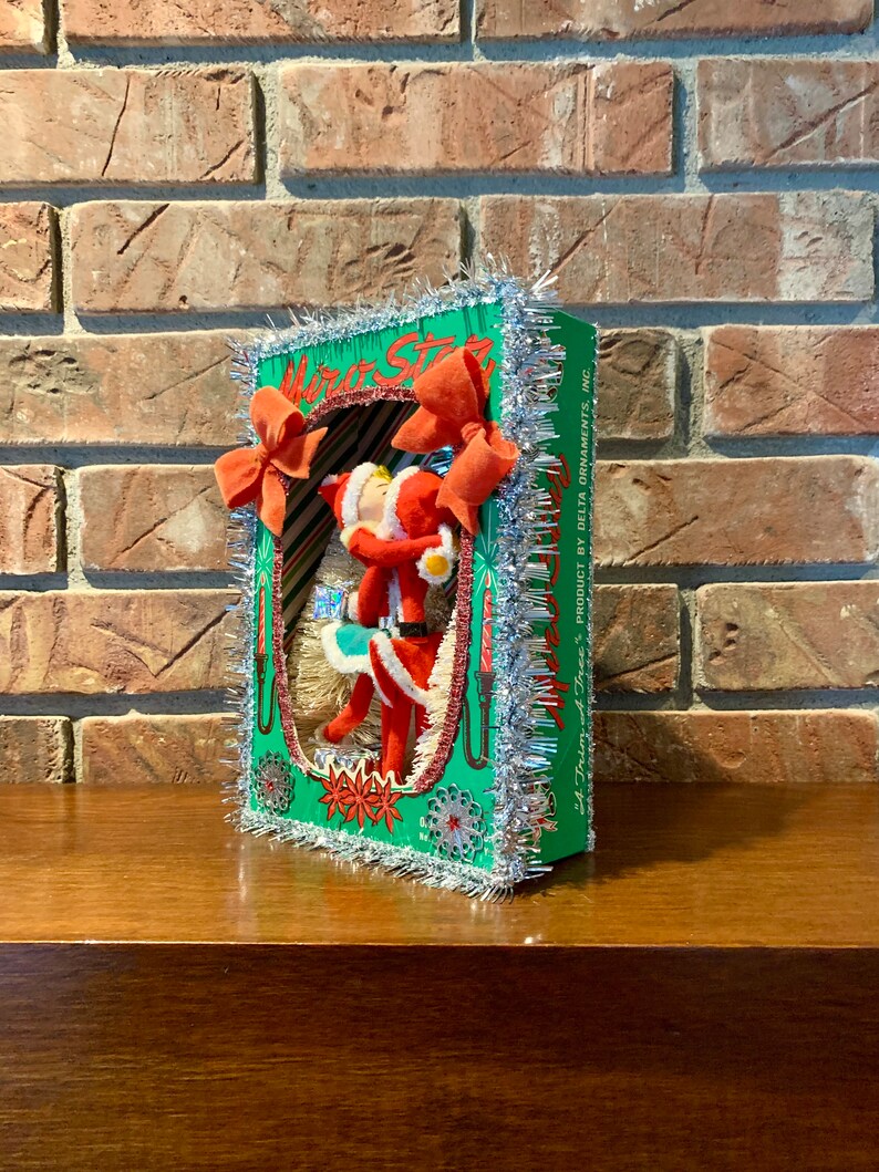 Op de afbeelding: Een vintage kerstboomversiering met een rood-witte kerstmanfiguur in een groen-witte doos met zilveren tinselrand. De doos heeft de tekst "Motor Star" en "A Trim of the Tree's Product by Delta Ornaments, Inc."