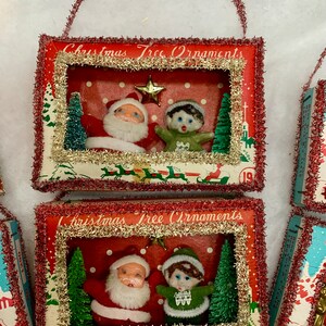 May include: Two vintage Christmas tree ornaments in the shape of small boxes with a red background and white polka dots. Each box has a window with a scene of Santa Claus and an elf inside. The boxes are decorated with red and gold tinsel and have the words "Christmas Tree Ornaments" printed on the front.