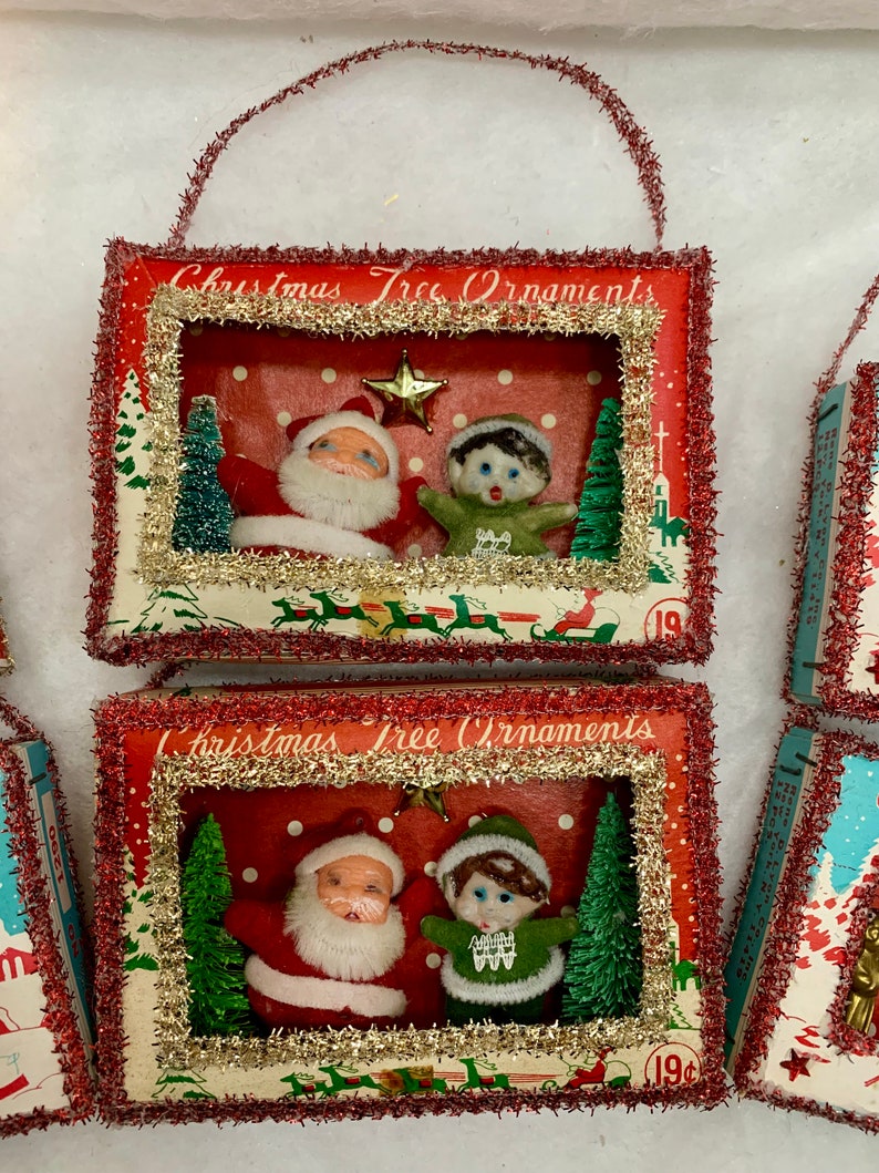 May include: Two vintage Christmas tree ornaments in the shape of small boxes with a red background and white polka dots. Each box has a window with a scene of Santa Claus and an elf inside. The boxes are decorated with red and gold tinsel and have the words "Christmas Tree Ornaments" printed on the front.