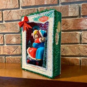 Op de afbeelding: Een vintage kerstboomversiering met een stel in een wintersc&egrave;ne. De versiering is gemaakt van glas en is versierd met glitter en tinsel. De doos is gelabeld met "Shiny Brite" en "American Made Glass Christmas Tree Ornaments".