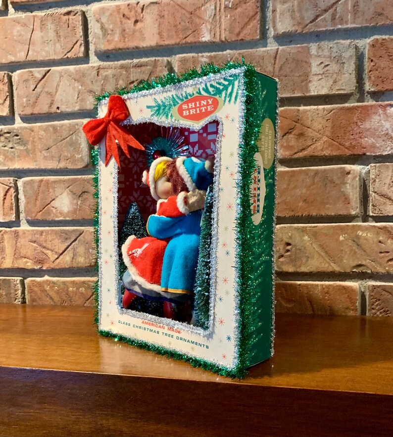 Op de afbeelding: Een vintage kerstboomversiering met een stel in een wintersc&egrave;ne. De versiering is gemaakt van glas en is versierd met glitter en tinsel. De doos is gelabeld met "Shiny Brite" en "American Made Glass Christmas Tree Ornaments".