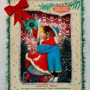 Op de afbeelding: Een vintage kerstboomversiering met een stel in rood en blauw gekleed dat elkaar kust. De versiering is ingelijst in een doos met een groene en zilveren rand. De doos heeft de tekst "Shiny Brite" en "American Made Glass Christmas Tree Ornaments".