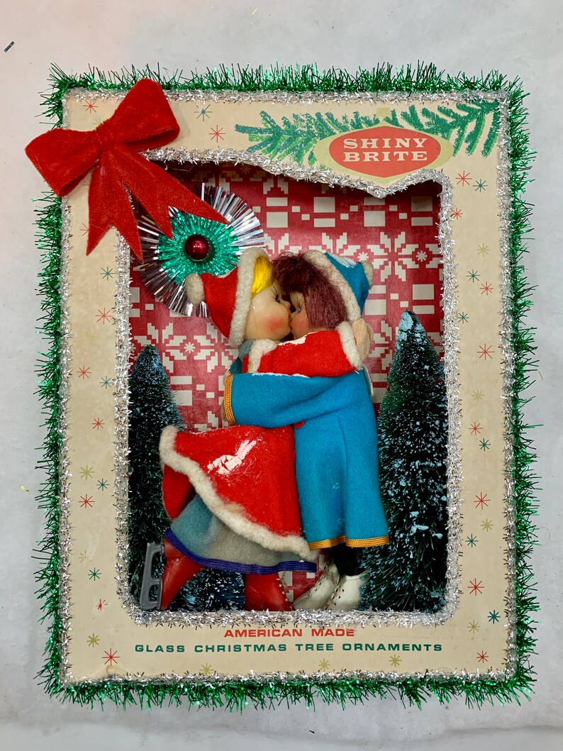 Op de afbeelding: Een vintage kerstboomversiering met een stel in rood en blauw gekleed dat elkaar kust. De versiering is ingelijst in een doos met een groene en zilveren rand. De doos heeft de tekst "Shiny Brite" en "American Made Glass Christmas Tree Ornaments".