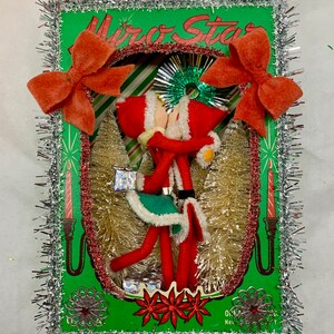 Op de afbeelding: Een vintage kerstdecoratie met twee rode vilten elven die elkaar kussen in een groen frame met een zilveren tinselrand. Het frame heeft de tekst "Mirror Star" erop gedrukt.