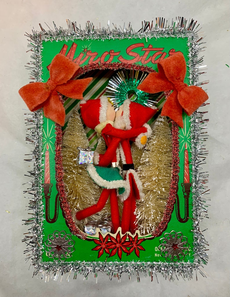 Op de afbeelding: Een vintage kerstdecoratie met twee rode vilten elven die elkaar kussen in een groen frame met een zilveren tinselrand. Het frame heeft de tekst "Mirror Star" erop gedrukt.