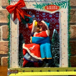 Op de afbeelding: Een vintage kerstsc&egrave;ne met twee poppen gekleed in rood en blauw, die elkaar kussen onder een glinsterende ster. De sc&egrave;ne is omlijst met groene tinsel en een rode strik. De tekst "Shiny Brite" is zichtbaar in de rechterbovenhoek.