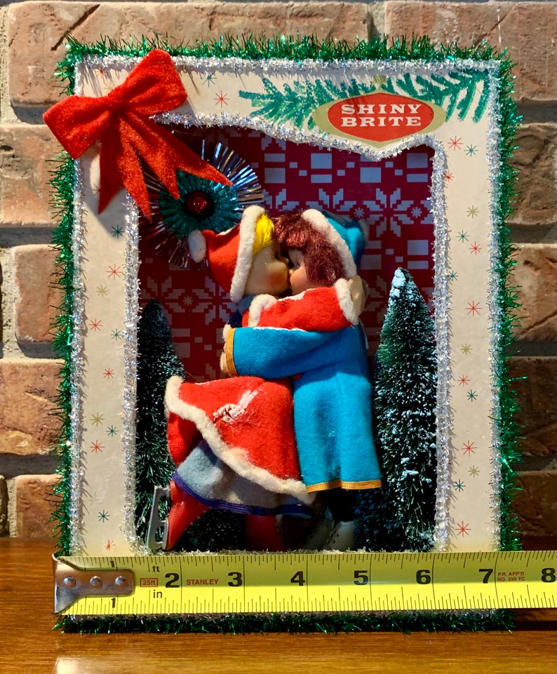 Op de afbeelding: Een vintage kerstsc&egrave;ne met twee poppen gekleed in rood en blauw, die elkaar kussen onder een glinsterende ster. De sc&egrave;ne is omlijst met groene tinsel en een rode strik. De tekst "Shiny Brite" is zichtbaar in de rechterbovenhoek.