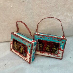 May include: Two vintage Christmas tree ornaments shaped like boxes with red and white trim. Each box has a window that shows a scene of angels and a Christmas tree. The boxes have a red and white tinsel trim and a loop for hanging.