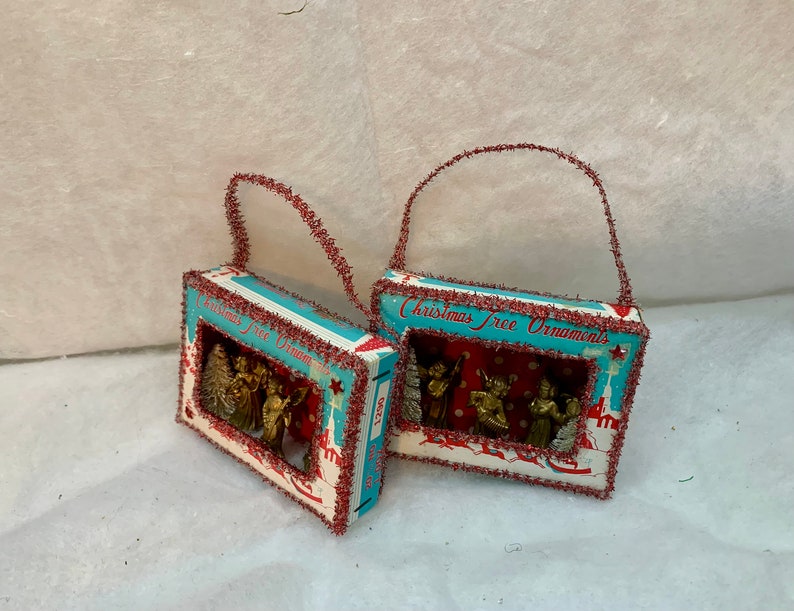 May include: Two vintage Christmas tree ornaments shaped like boxes with red and white trim. Each box has a window that shows a scene of angels and a Christmas tree. The boxes have a red and white tinsel trim and a loop for hanging.