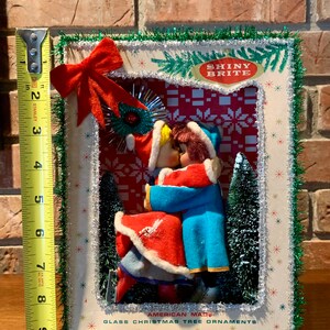 Op de afbeelding: Een vintage kerstboomversieringset met een stel in een besneeuwde sc&egrave;ne. De versiering is omlijst met groene tinsel en heeft de tekst "Shiny Brite" en "American Made Glass Christmas Tree Ornaments".