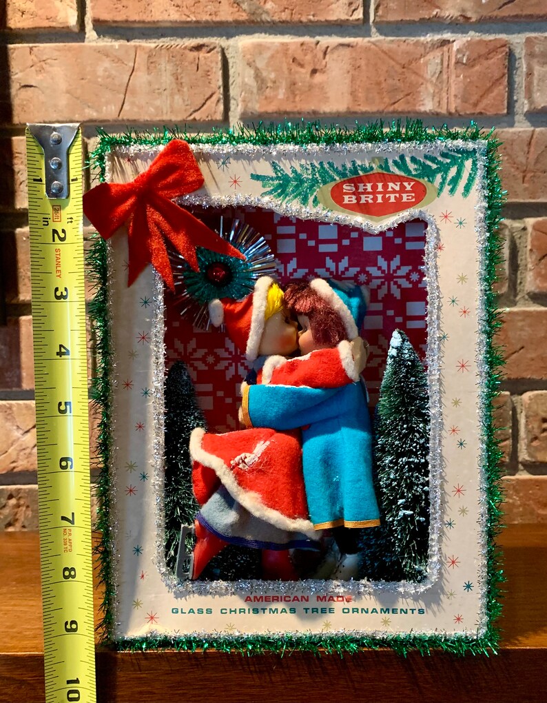 Op de afbeelding: Een vintage kerstboomversieringset met een stel in een besneeuwde sc&egrave;ne. De versiering is omlijst met groene tinsel en heeft de tekst "Shiny Brite" en "American Made Glass Christmas Tree Ornaments".