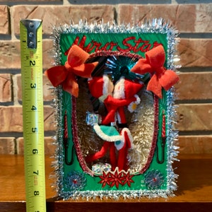 Op de afbeelding: Vintage kerstversiering met twee rode vilten elven die hand in hand staan in een groene en rode sc&egrave;ne met een zilveren tinselrand. De sc&egrave;ne bevat een rode kaars, een rode strik en een rode ster. De tekst "Merry Star" is zichtbaar bovenaan de sc&egrave;ne.
