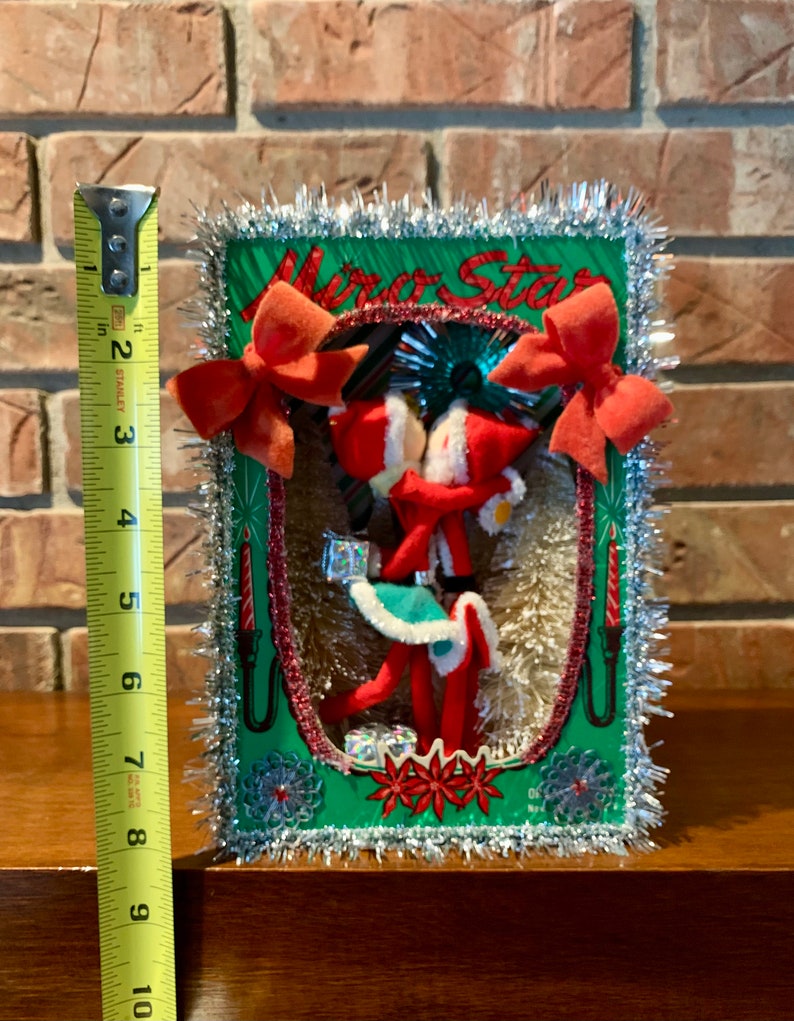 Op de afbeelding: Vintage kerstversiering met twee rode vilten elven die hand in hand staan in een groene en rode sc&egrave;ne met een zilveren tinselrand. De sc&egrave;ne bevat een rode kaars, een rode strik en een rode ster. De tekst "Merry Star" is zichtbaar bovenaan de sc&egrave;ne.