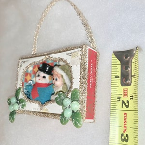 May include: A vintage Christmas ornament made from a matchbox with a picture of a snowman and a girl inside. The ornament is decorated with silver tinsel and green holly leaves with white berries. The matchbox is labeled "Shiny White".