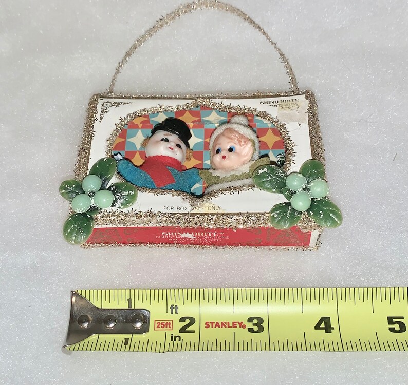 May include: A vintage Christmas ornament featuring two small dolls, a snowman and a girl, in a framed scene. The ornament is made of paper mache and has a gold glitter border. The ornament is approximately 2 inches tall.