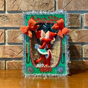 Op de afbeelding: Een vintage kerstversiering met een stel vilten elven die elkaar kussen in een groen frame met zilveren tinselrand. Het frame heeft de tekst "Miro Star" erop gedrukt.