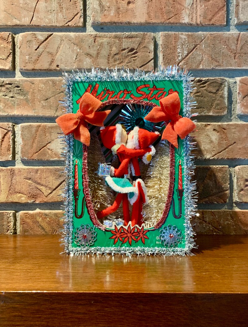 Op de afbeelding: Een vintage kerstversiering met een stel vilten elven die elkaar kussen in een groen frame met zilveren tinselrand. Het frame heeft de tekst "Miro Star" erop gedrukt.