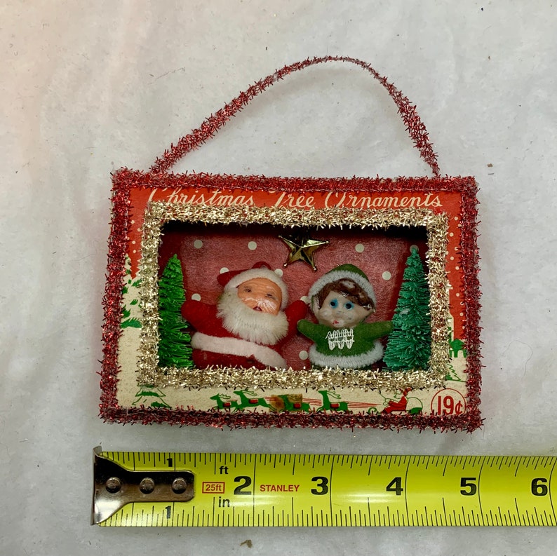 May include: Vintage Christmas tree ornament featuring a red and white Santa Claus and a green elf in a snowy scene. The ornament is framed in red and gold glitter and has a red and white string hanger. The text "Christmas Tree Ornaments" is printed on the top of the ornament. The price tag reads "19c".