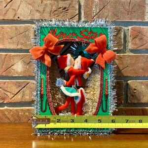 Op de afbeelding: Een vintage kerstversiering met een groene kaart waarop de woorden "Mira Star" in rood-witte letters staan. De kaart is versierd met rode strikken, zilveren tinsel en twee rood-witte kerstmanfiguren die een cadeaudoos vasthouden. De versiering is ongeveer 18 cm hoog.