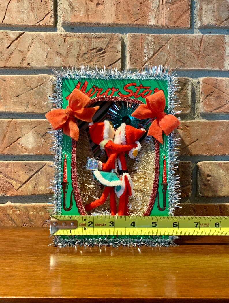 Op de afbeelding: Een vintage kerstversiering met een groene kaart waarop de woorden "Mira Star" in rood-witte letters staan. De kaart is versierd met rode strikken, zilveren tinsel en twee rood-witte kerstmanfiguren die een cadeaudoos vasthouden. De versiering is ongeveer 18 cm hoog.